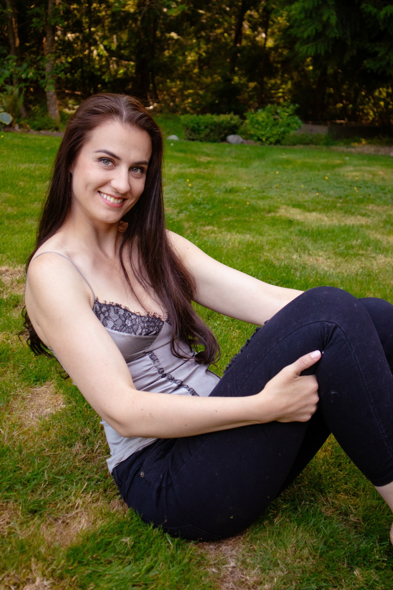Young woman with long dark hair sitting on grass in a park, smiling at the camera.