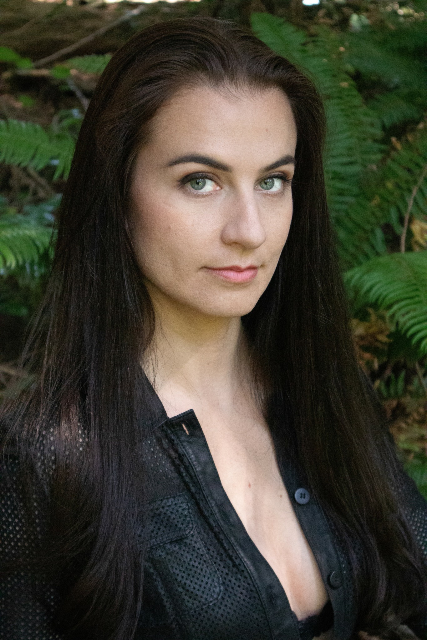 Young woman with long dark hair and blue eyes outdoors among green plants.