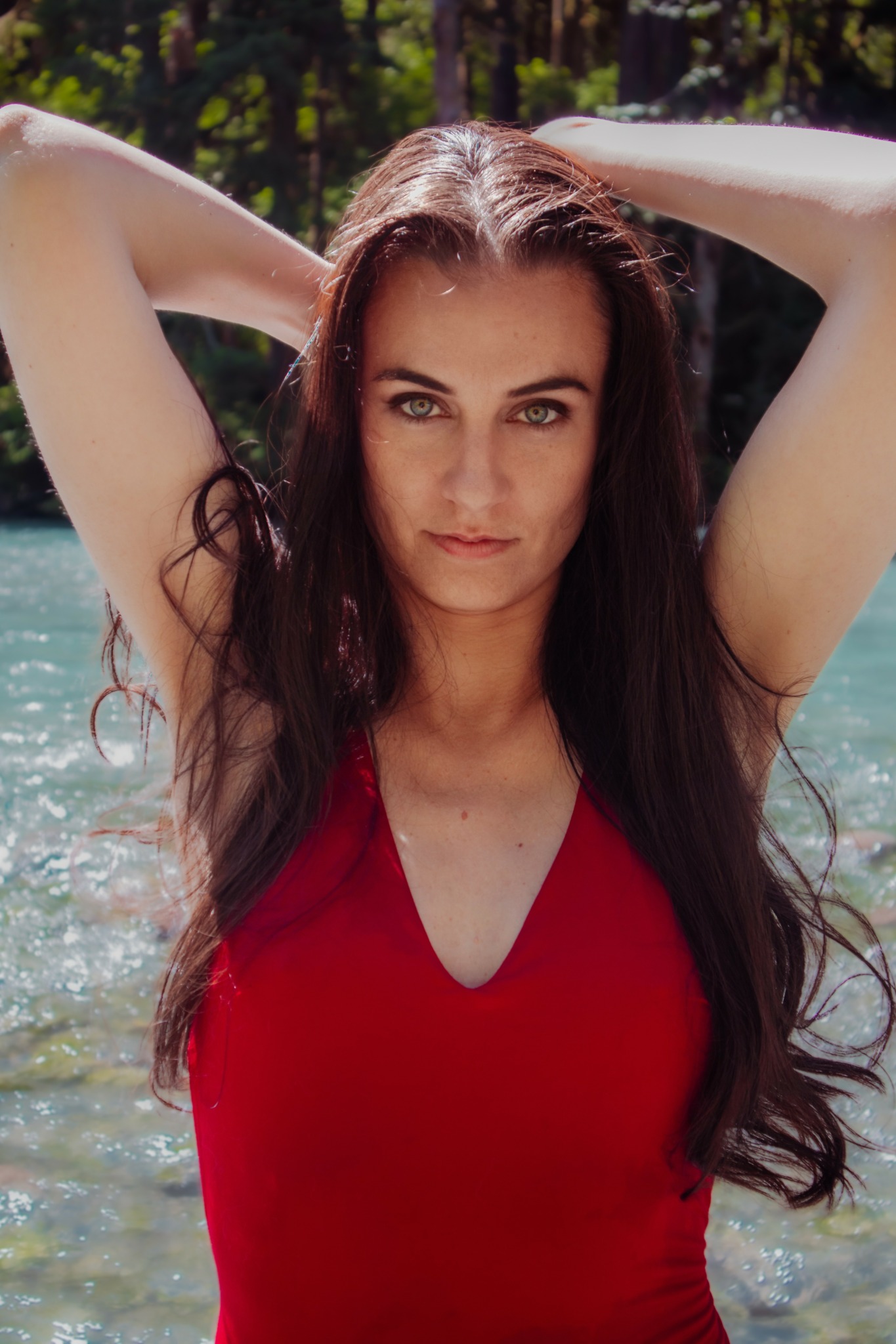 Woman with long dark hair in a red swimsuit, arms raised, outdoors near water and trees.