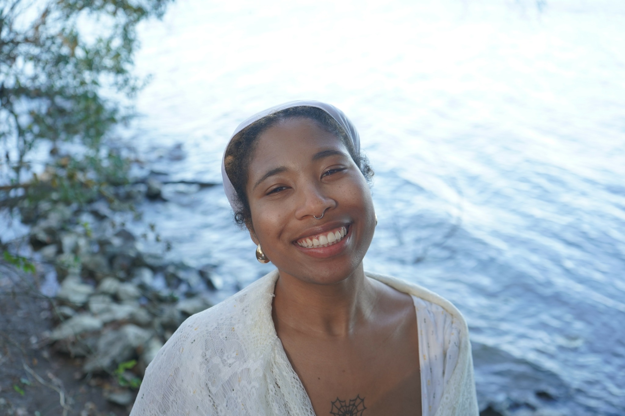 Smiling woman with dark hair tied back, wearing a light-colored top, standing near water with trees in the background.