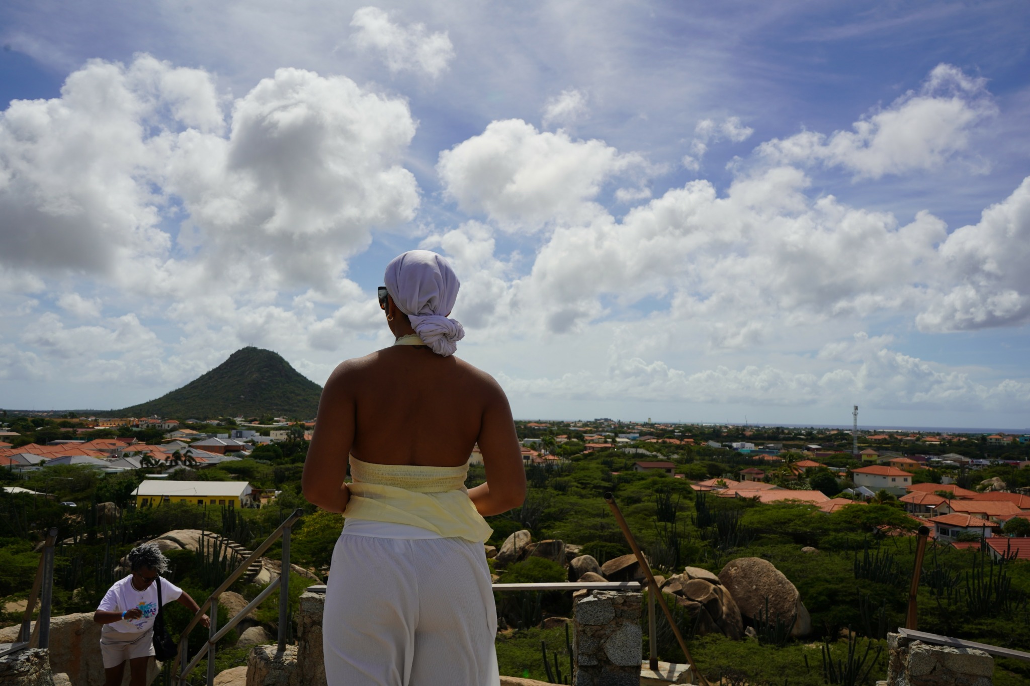 Person with a white head covering and yellow top looking over a town with a mountain in the background.