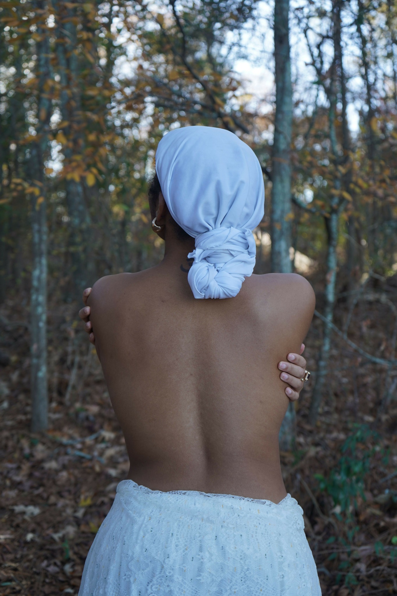 Person with a towel around waist and a white cloth on head, standing outdoors among trees.