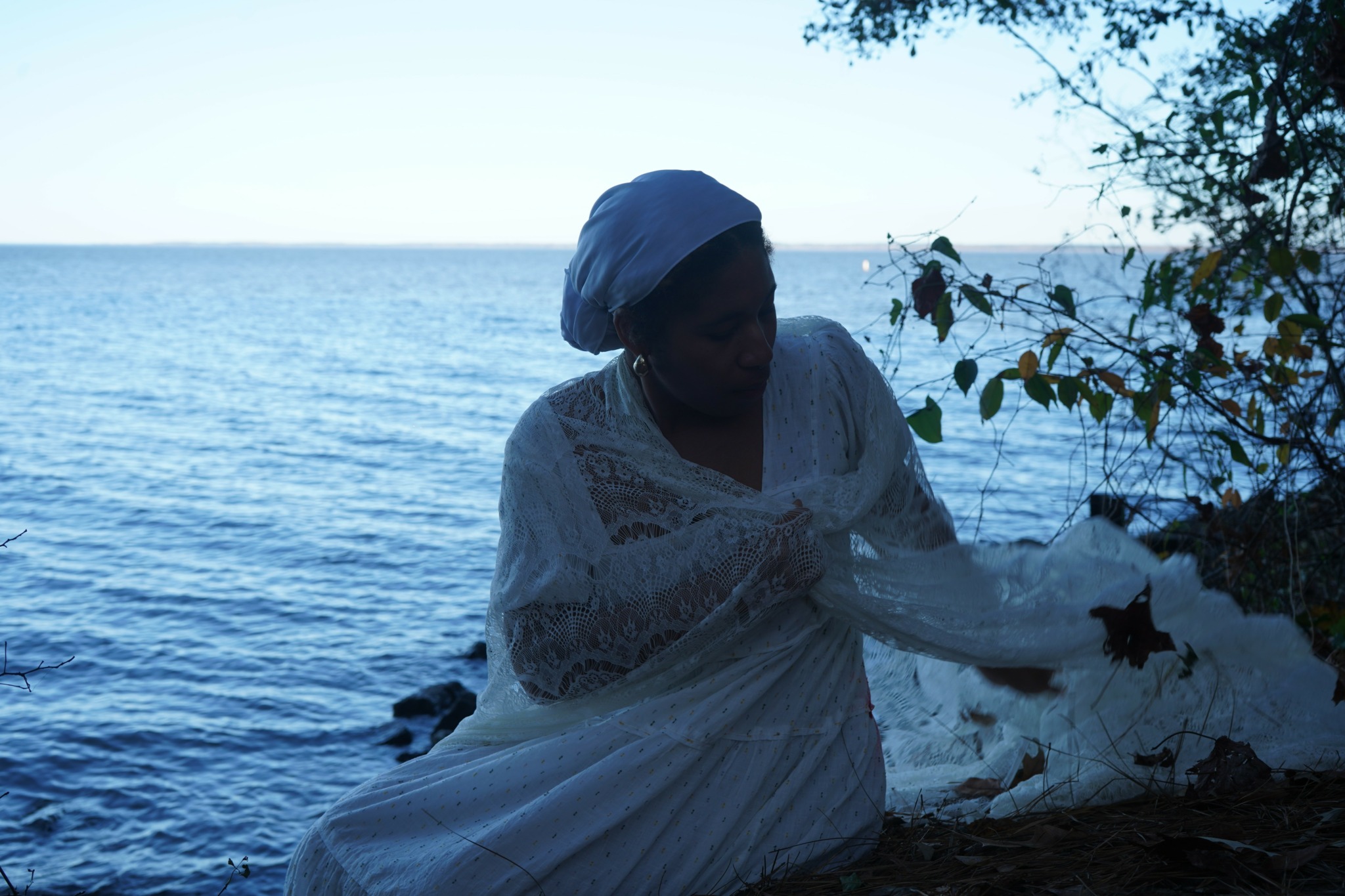 Person in white dress and head covering sitting by water near trees, facing downward, with water and sky in background.
