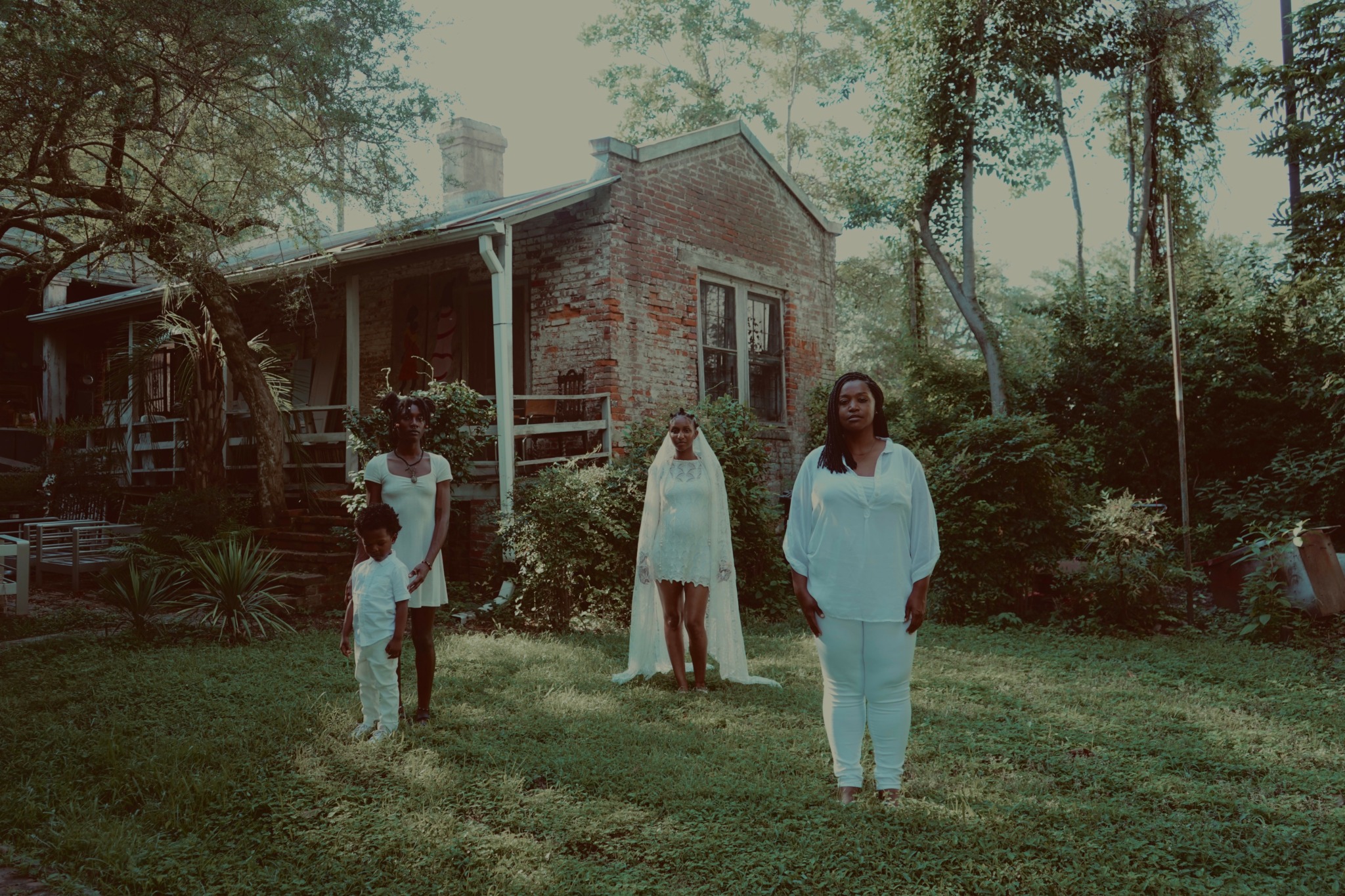 Four people standing on a grassy yard in front of a brick house with trees and bushes around.