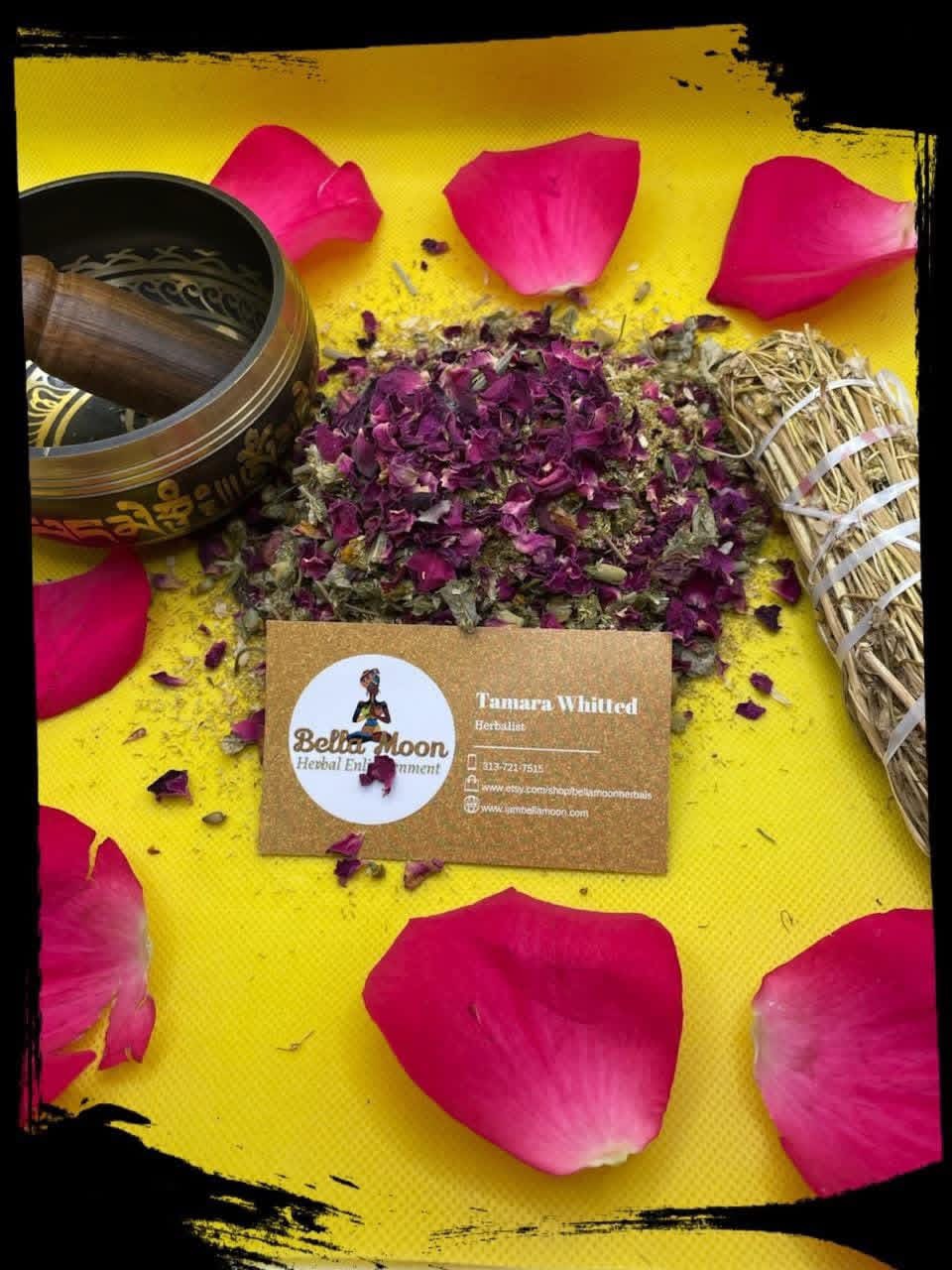 Dried purple flowers, pink rose petals, a business card, and a rolled bundle on a yellow surface.