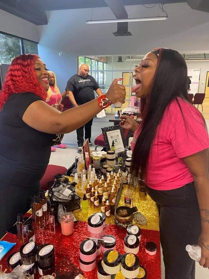 Two women smiling and sharing makeup products at a table indoors, with other people in the background.