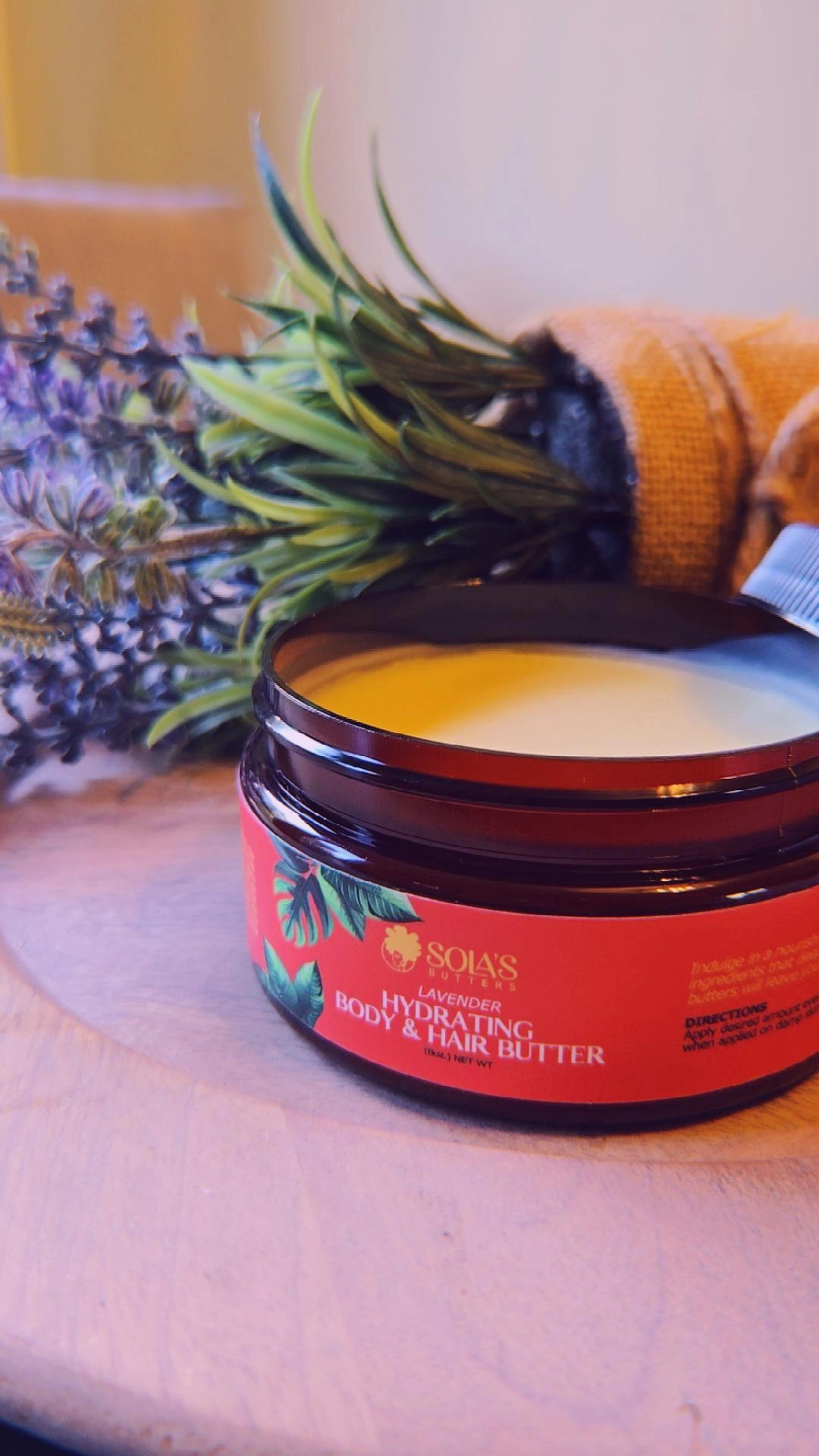 Open jar of body butter with lavender and green leaves nearby on wooden surface.