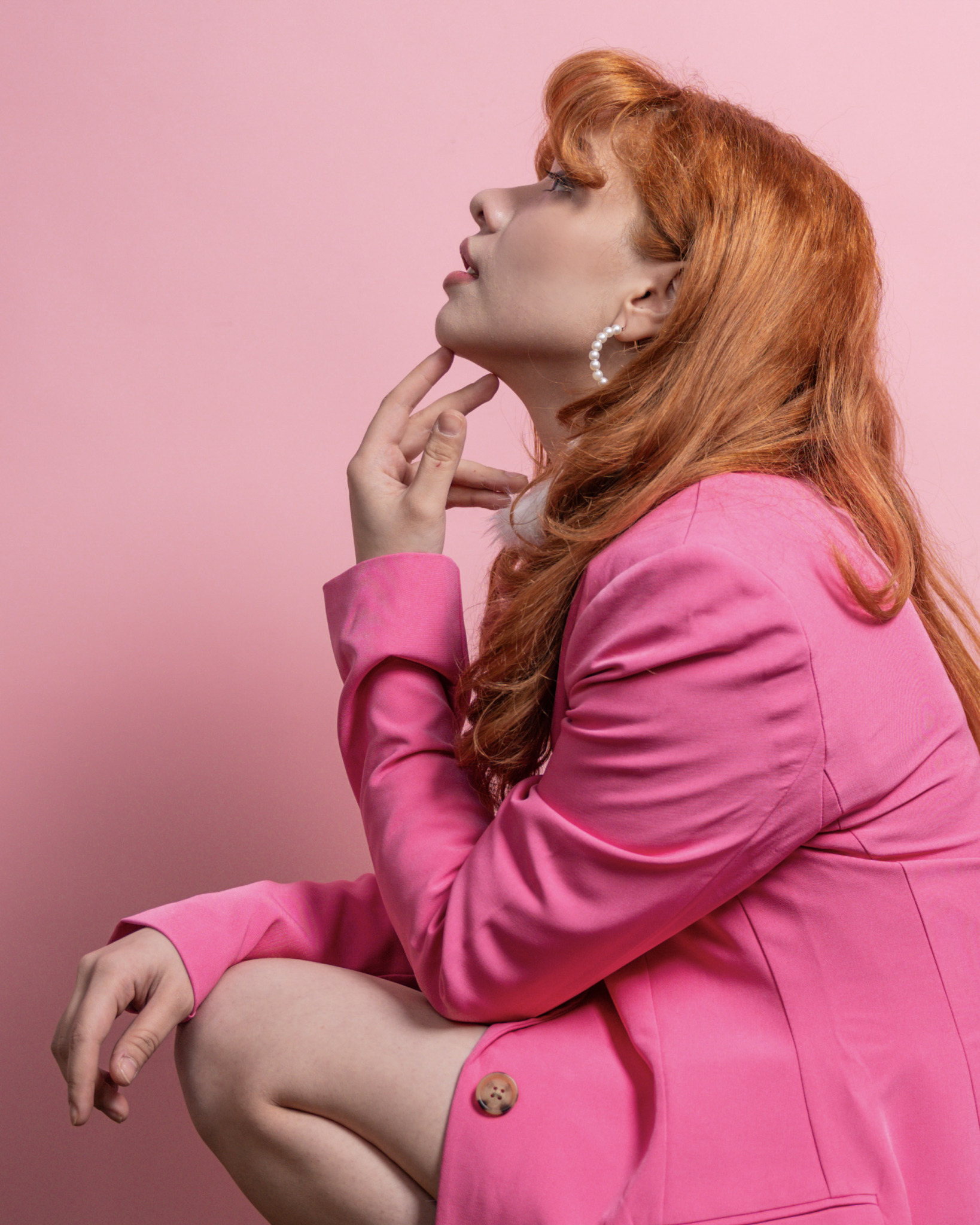 Side profile of a woman with red hair, wearing a pink blazer and pearl earrings, against a pink background.