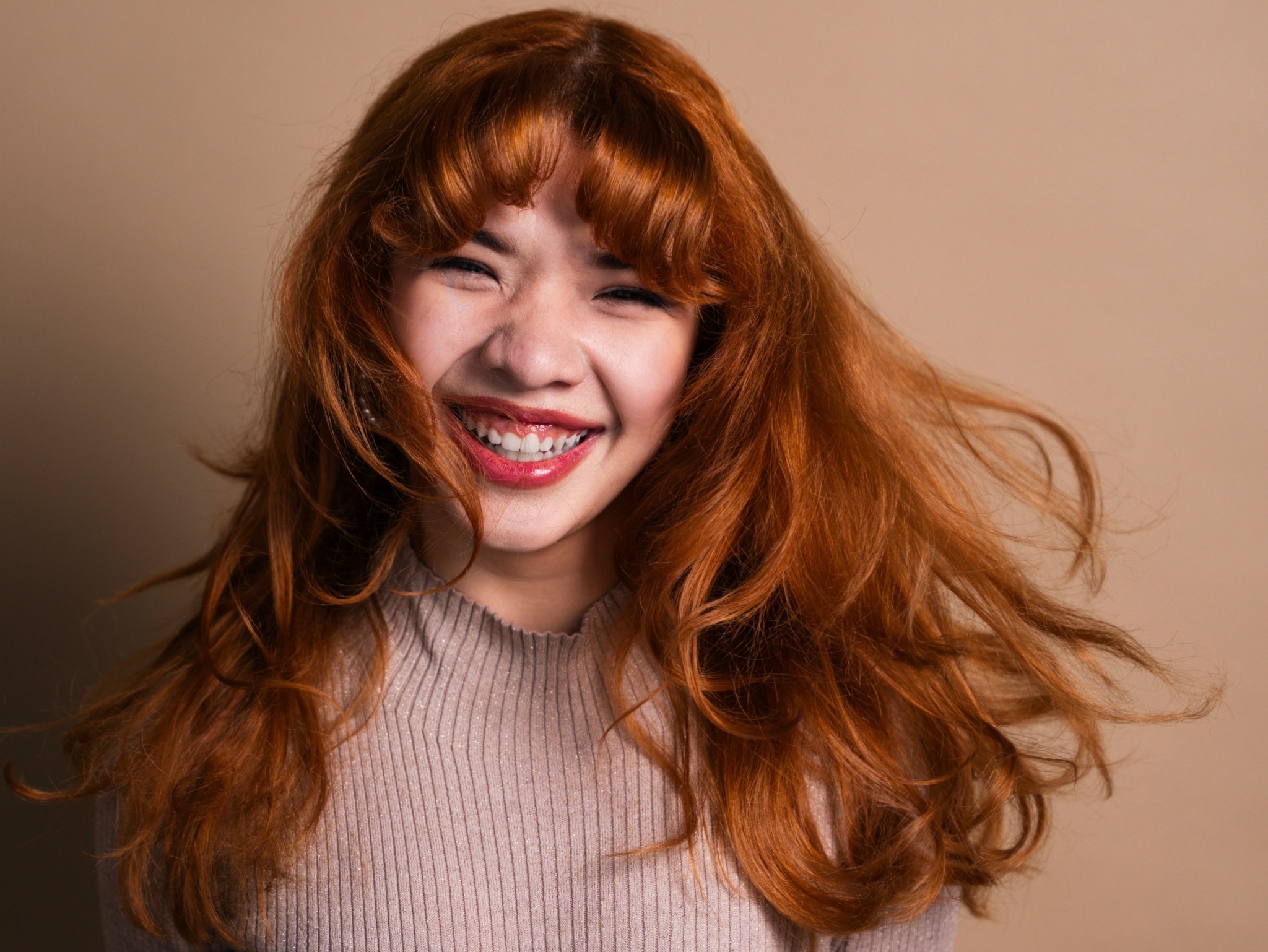 Young woman with long, wavy red hair smiling and winking, wearing a beige top, against a plain background.