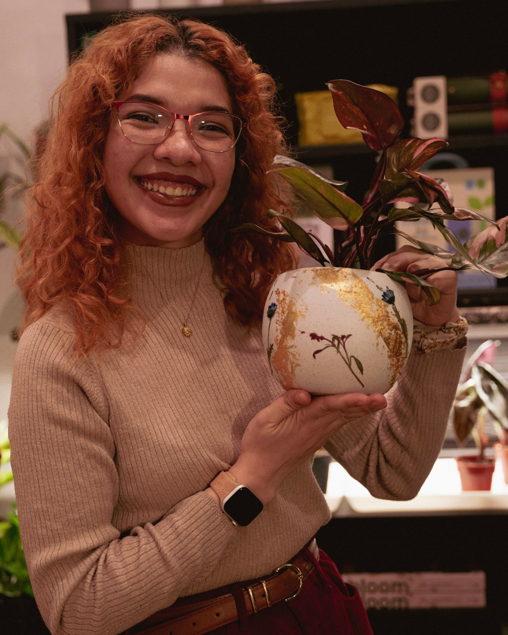 Smiling woman with curly red hair and glasses holding a potted plant in a decorative vase.