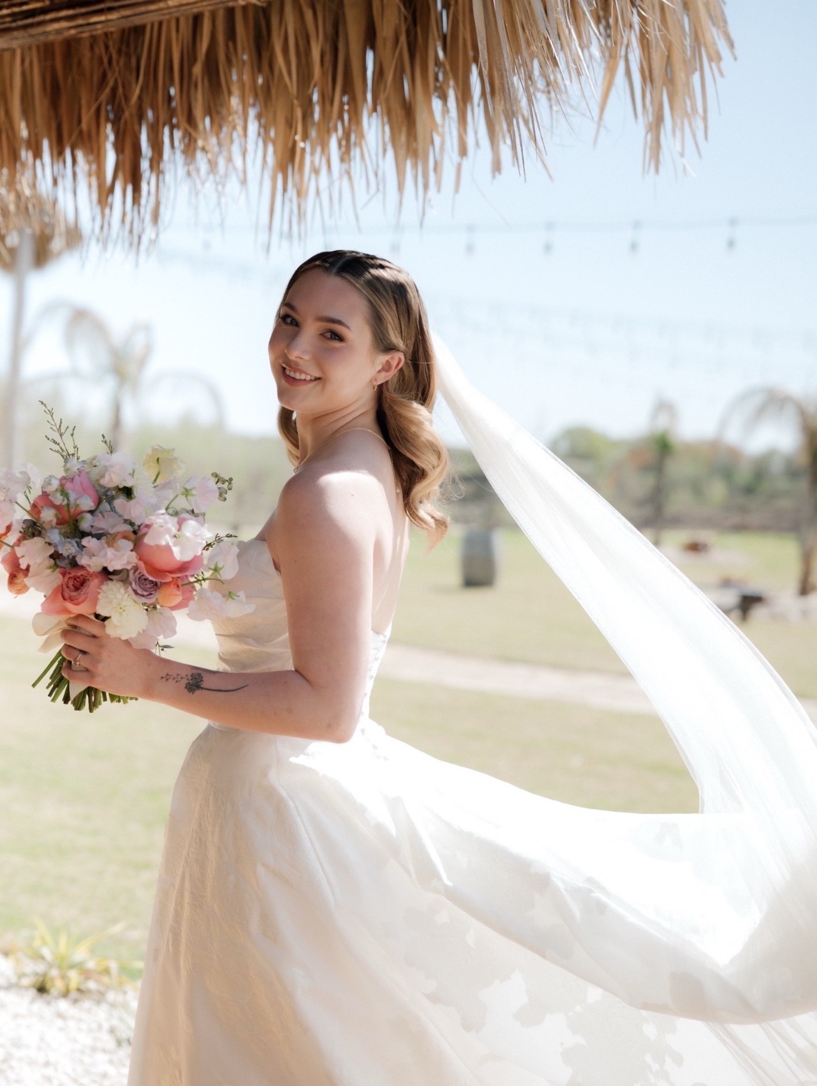 Young woman in wedding dress holding a bouquet, smiling outdoors under a thatched roof.