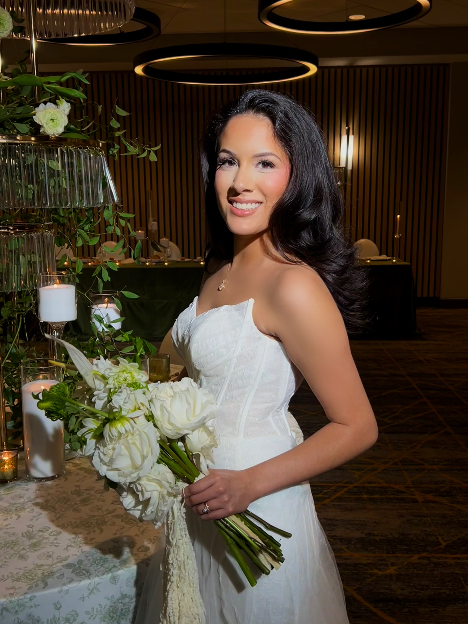 Woman in white dress holding a bouquet of white flowers indoors with dark wood walls and circular ceiling lights.