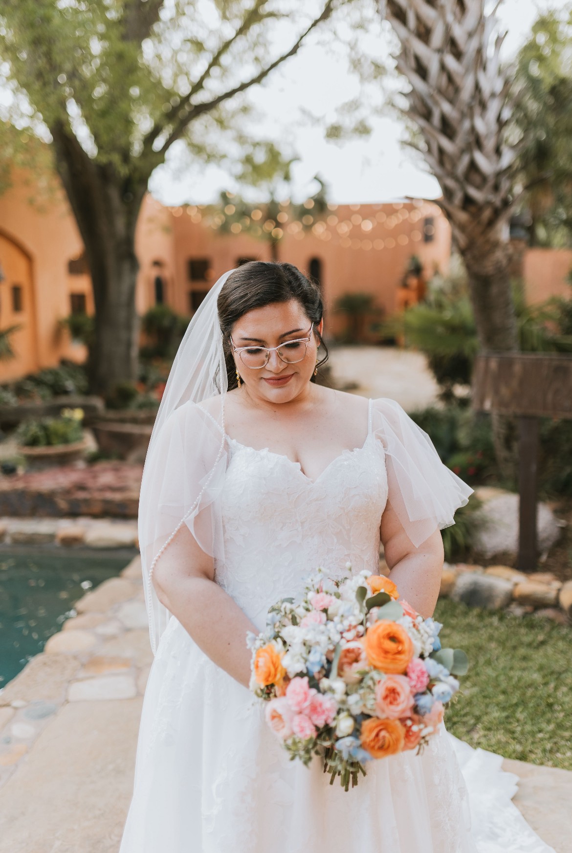 Bride in wedding dress holding a colorful bouquet outdoors with trees and buildings in background.