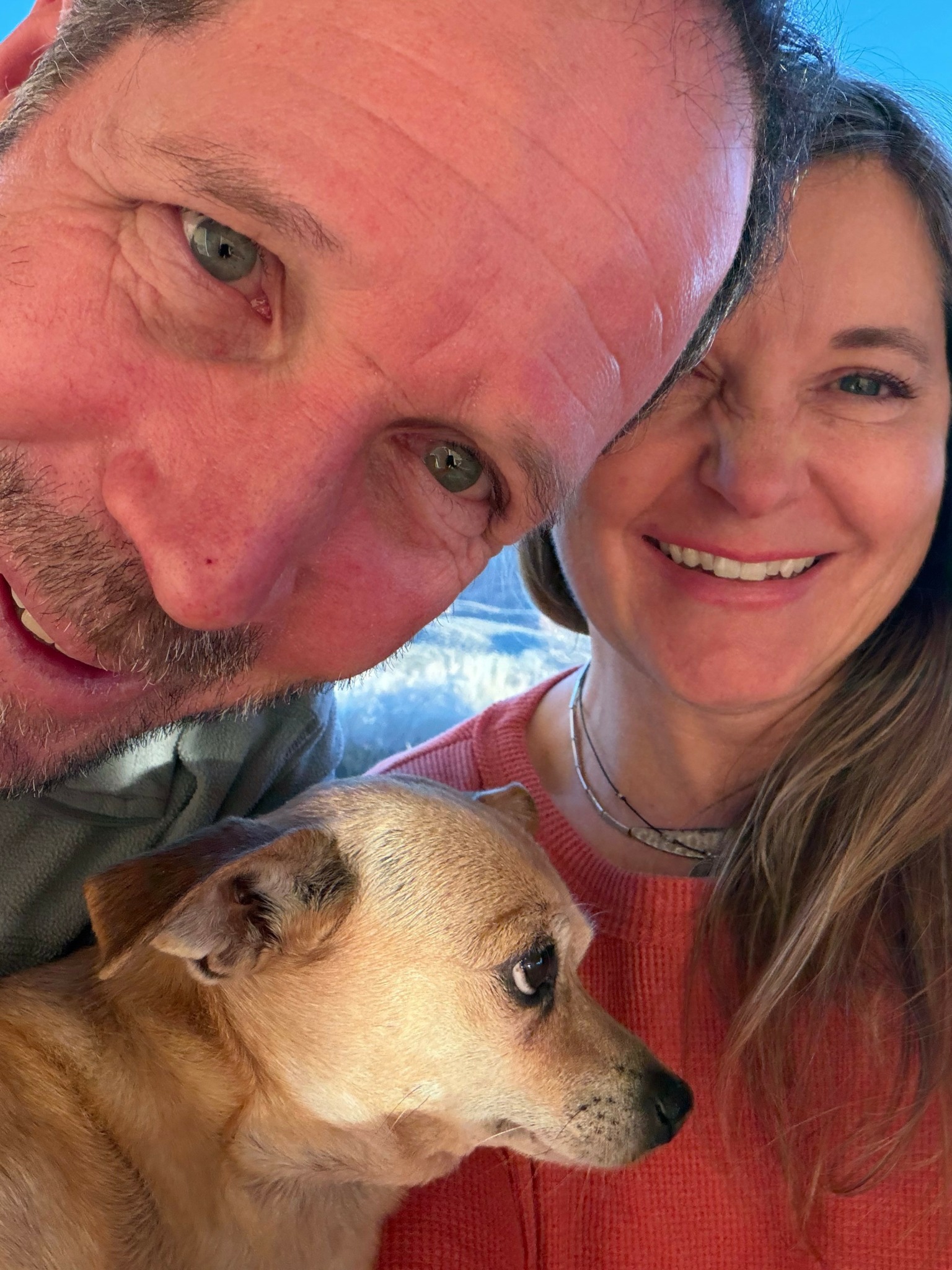 Close-up of two smiling people and a dog outdoors, background of blue sky and clouds.