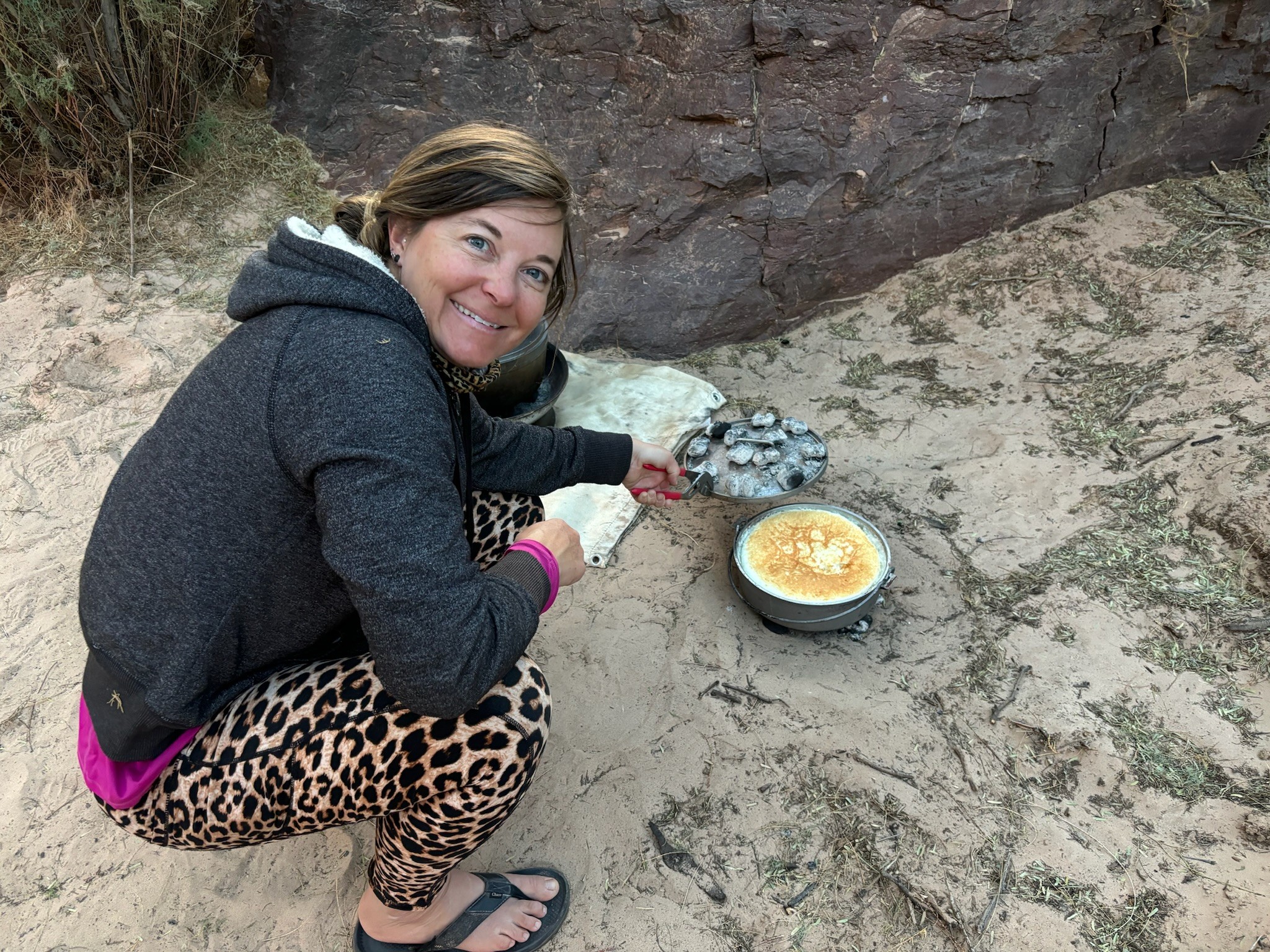 Woman crouching outdoors, smiling, holding a rock near a bowl of food on sandy ground.