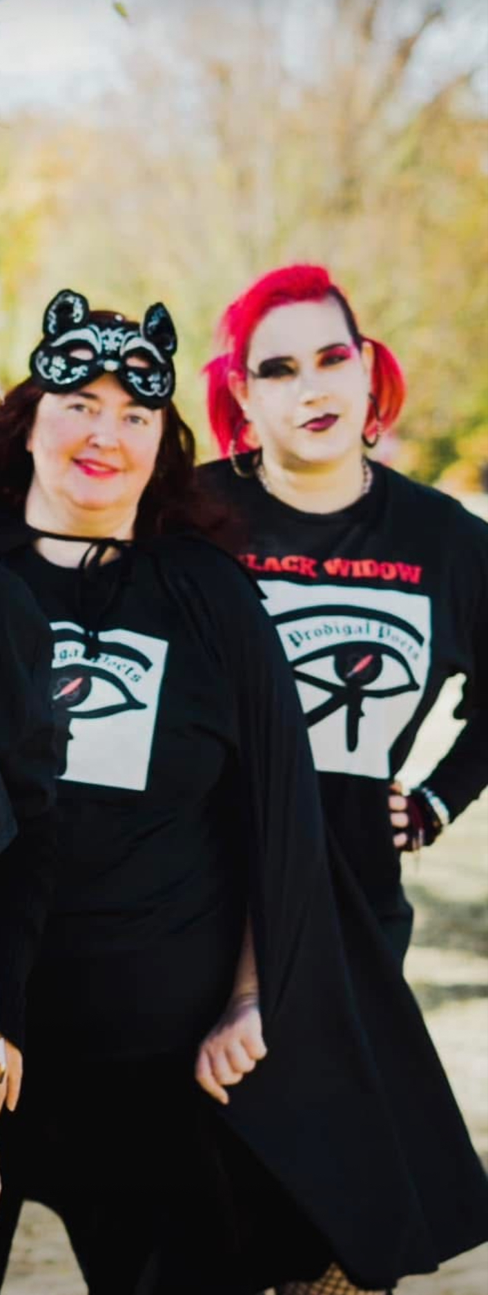 Two women standing outdoors, one with black hair and bear-shaped glasses, the other with bright pink hair, both wearing black clothing.