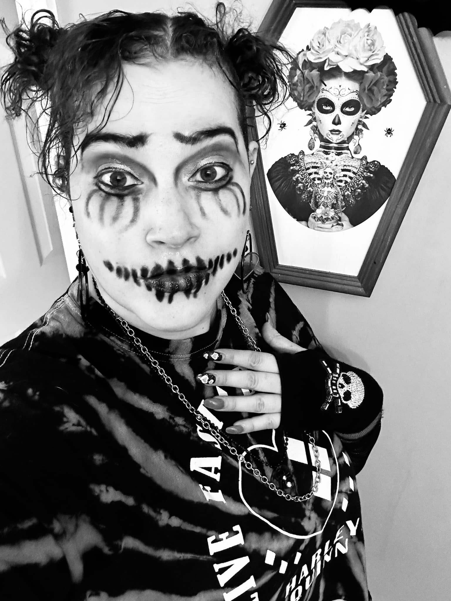 Person with face paint resembling a skeleton, curly hair, and dark clothing, standing in front of a framed artwork of a woman.