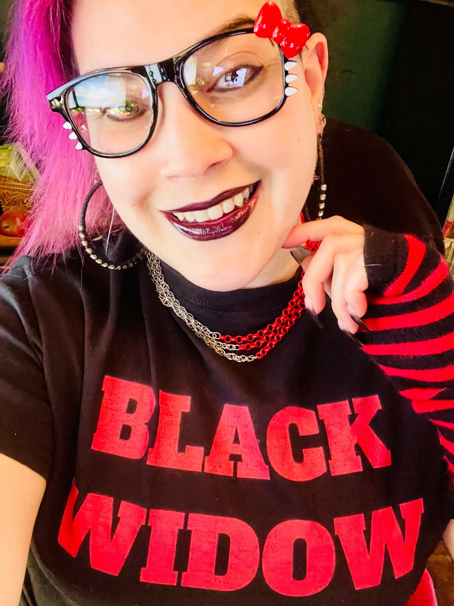 Person with pink hair, glasses, dark lipstick, wearing a black shirt with red text, and jewelry, smiling.