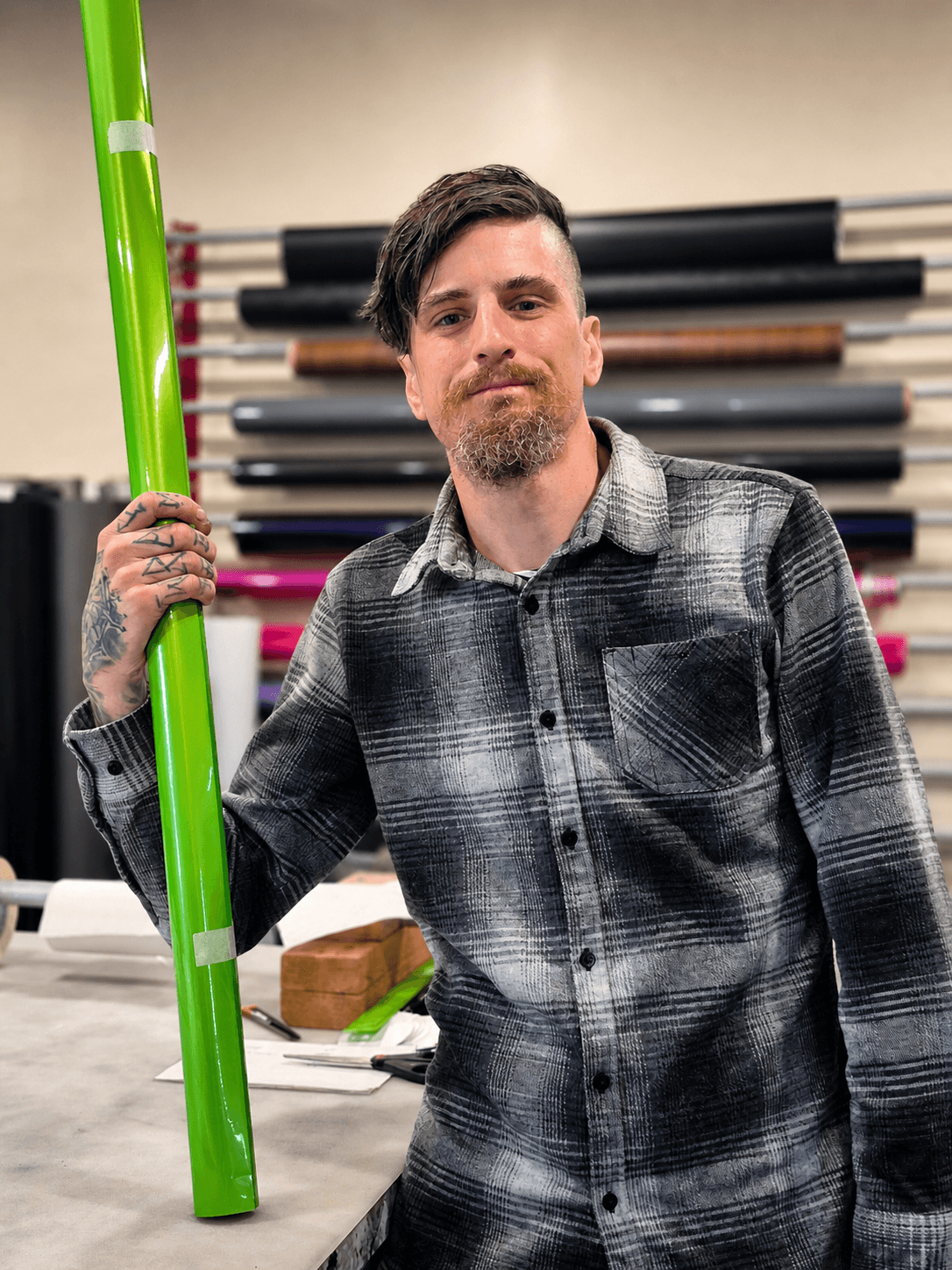 Man with beard and tattoos holding a green pole in a room with stacked black and pink mats.