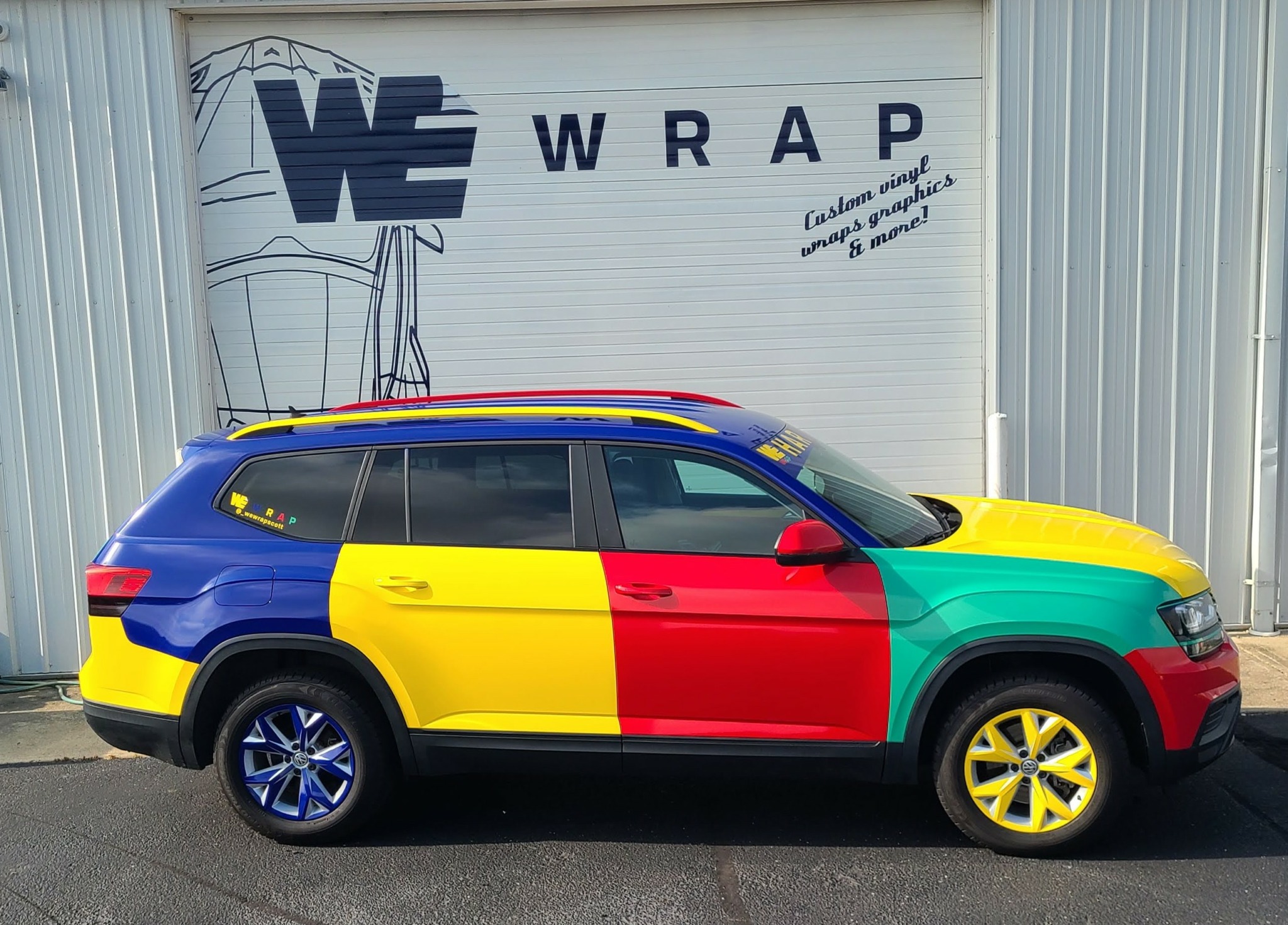 Colorful car with yellow, red, green, and blue sections parked outside a building with a sign that says 'WE WRAP'.