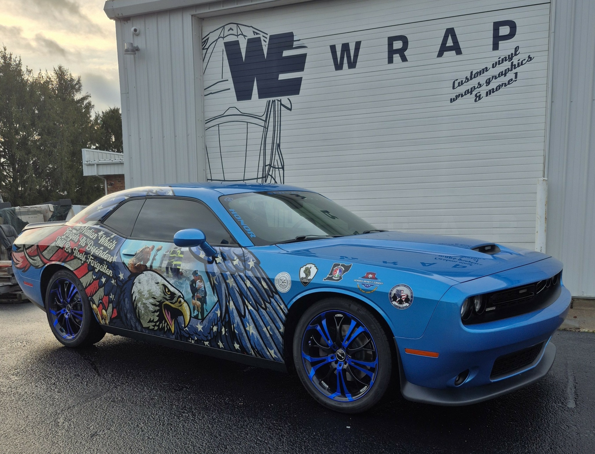 Blue sports car with eagle and graphic design parked outside a building with a sign reading 'WE WRAP'.