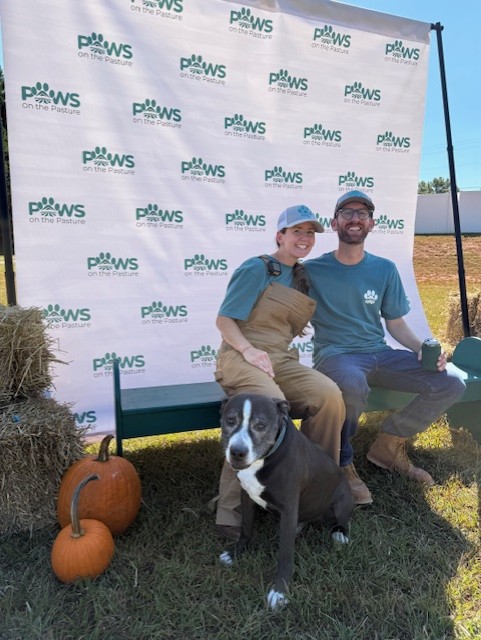Two people sitting on a bench with a black and white dog in front, outdoors with pumpkins nearby and a backdrop with logos.