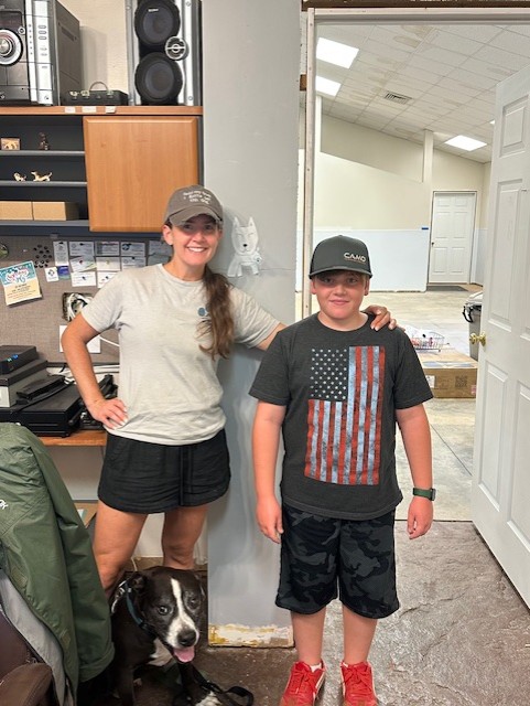 A woman and a boy stand indoors, smiling, with a dog sitting nearby. The woman has her arm around the boy.