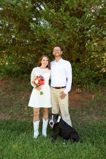 A smiling woman in a white dress holding a bouquet of flowers, standing next to a man in a white shirt and beige pants, with a black and white dog sitting in front of them, outdoors with green trees.