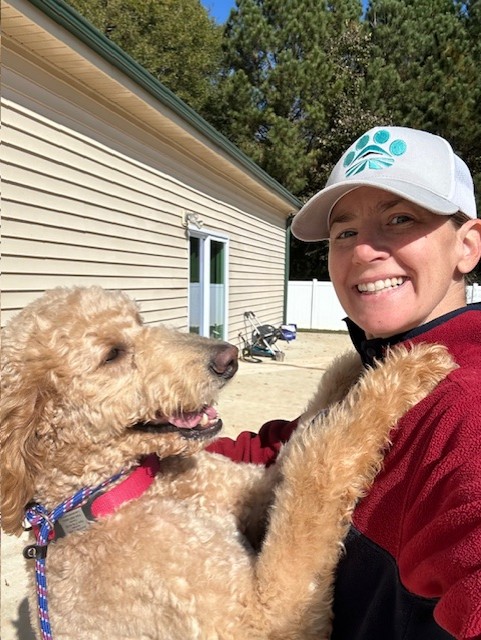 Person smiling with a dog outdoors near a house, trees in background.