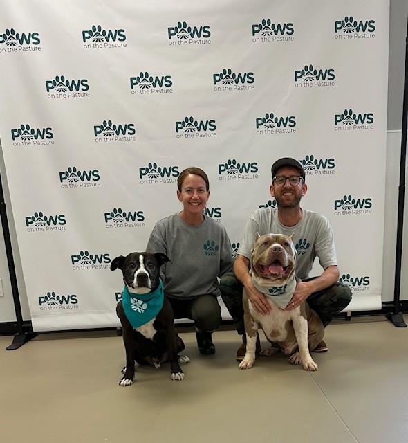 Two people kneel with two dogs in front of a Paw's on the Future backdrop.