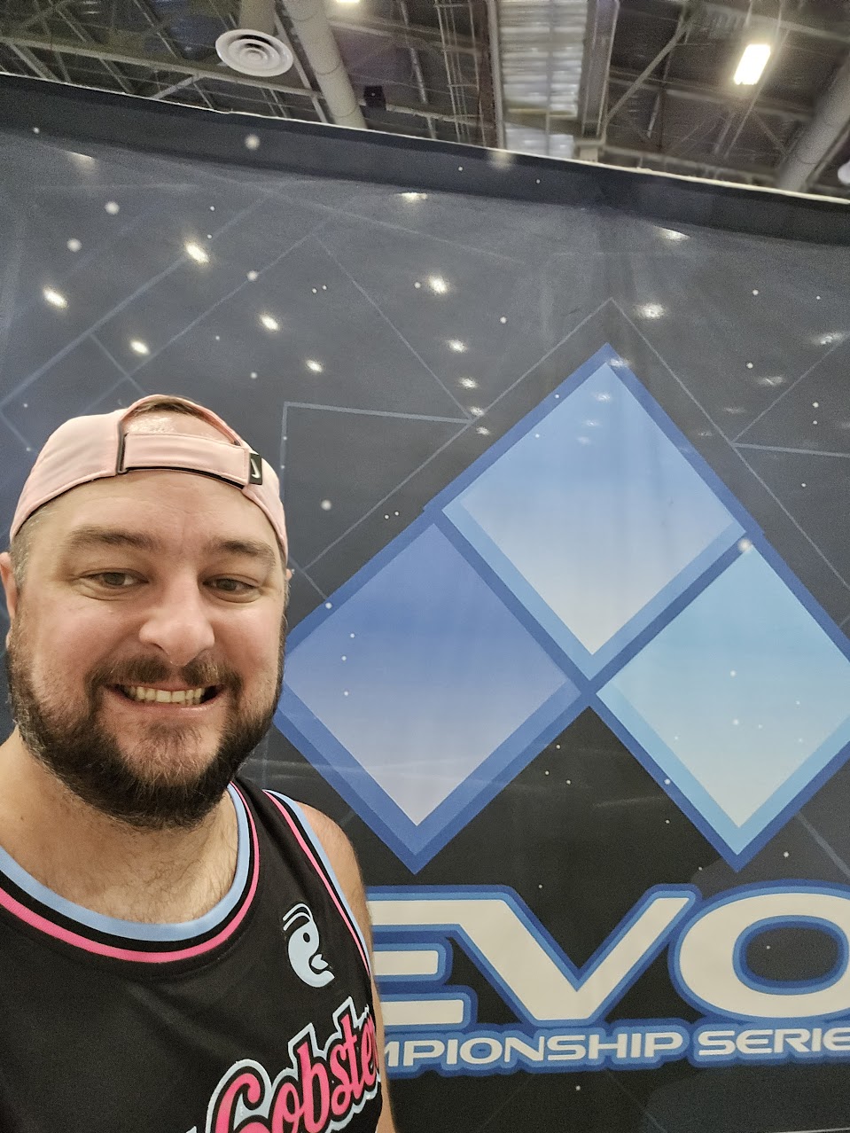 Smiling man with beard wearing a pink cap backwards and a black sports jersey, standing in front of a large blue geometric logo.