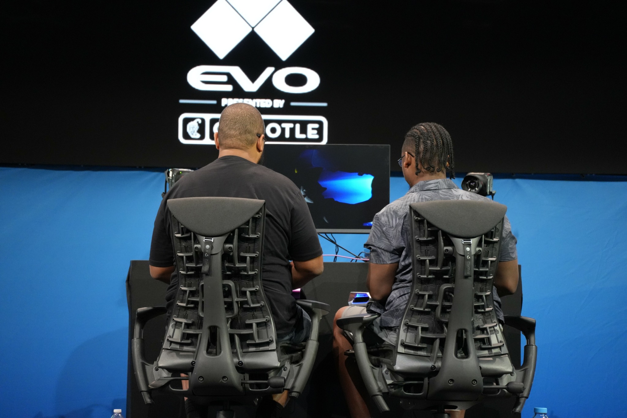 Two people sit in gaming chairs facing a large screen with the EVO logo and text, in a dark room.