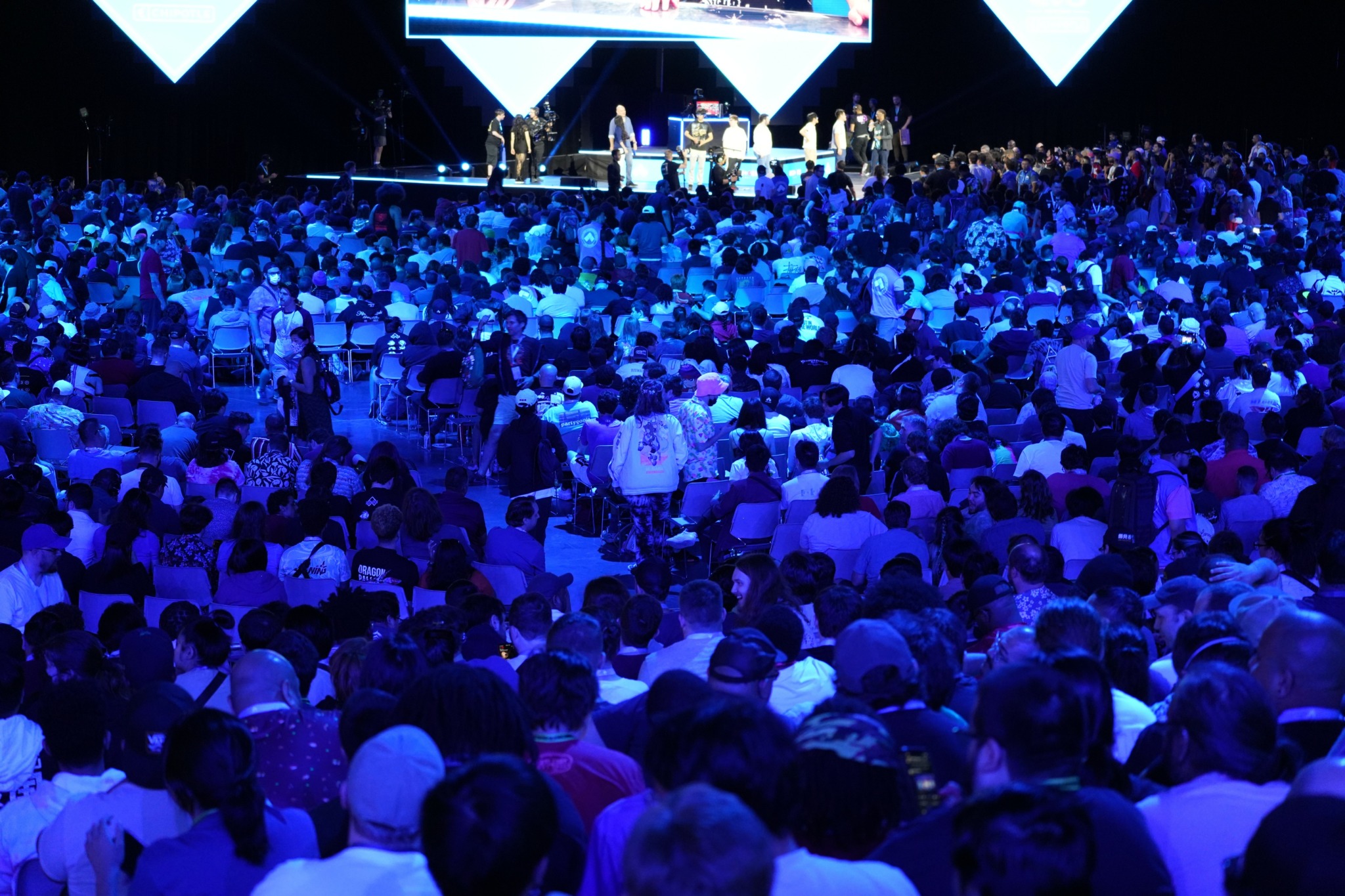 Large audience seated facing stage with performers and screens, illuminated in blue lighting.
