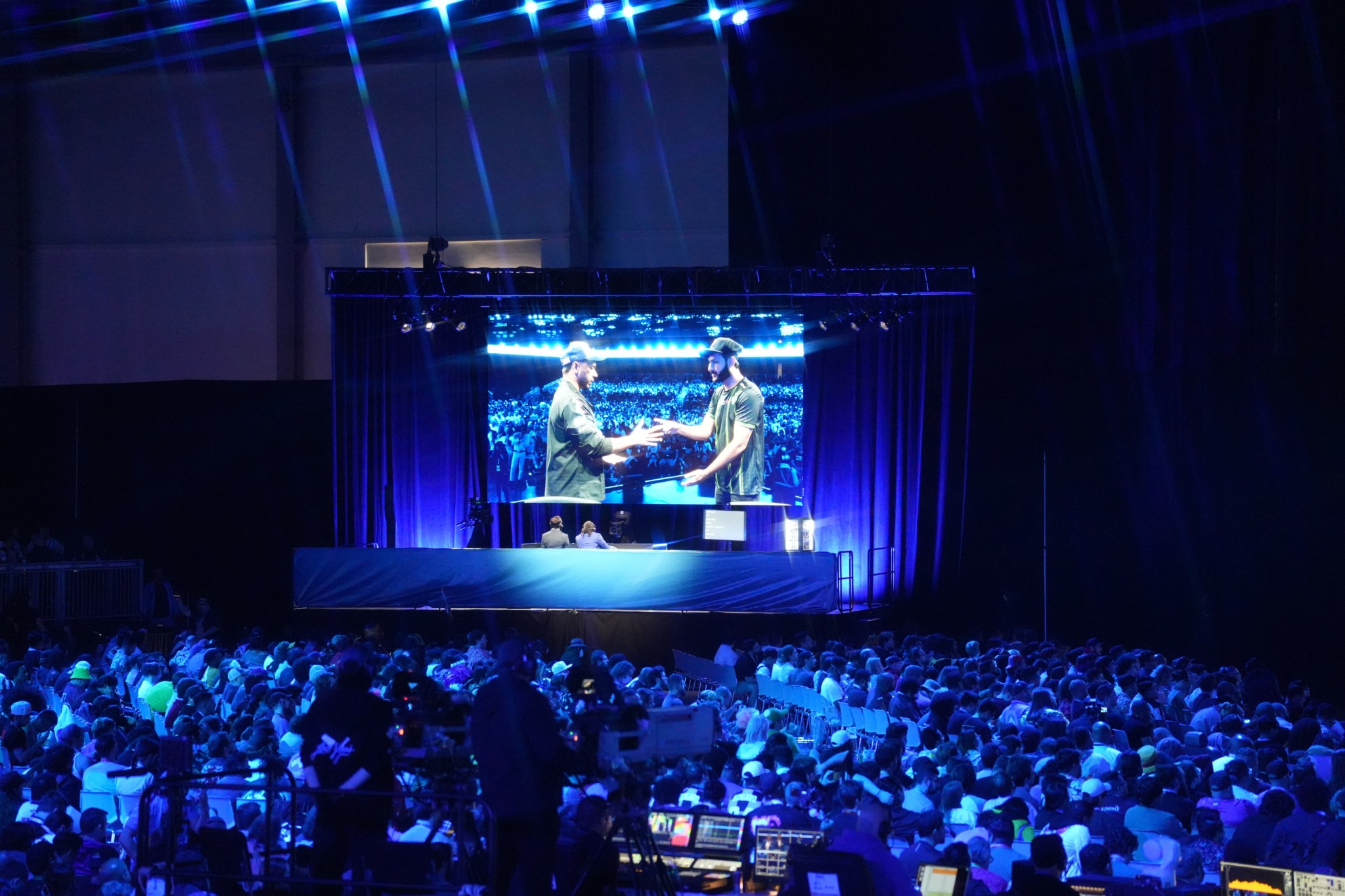 Stage with two people shaking hands, large audience seated, blue lighting, and a screen displaying the scene.