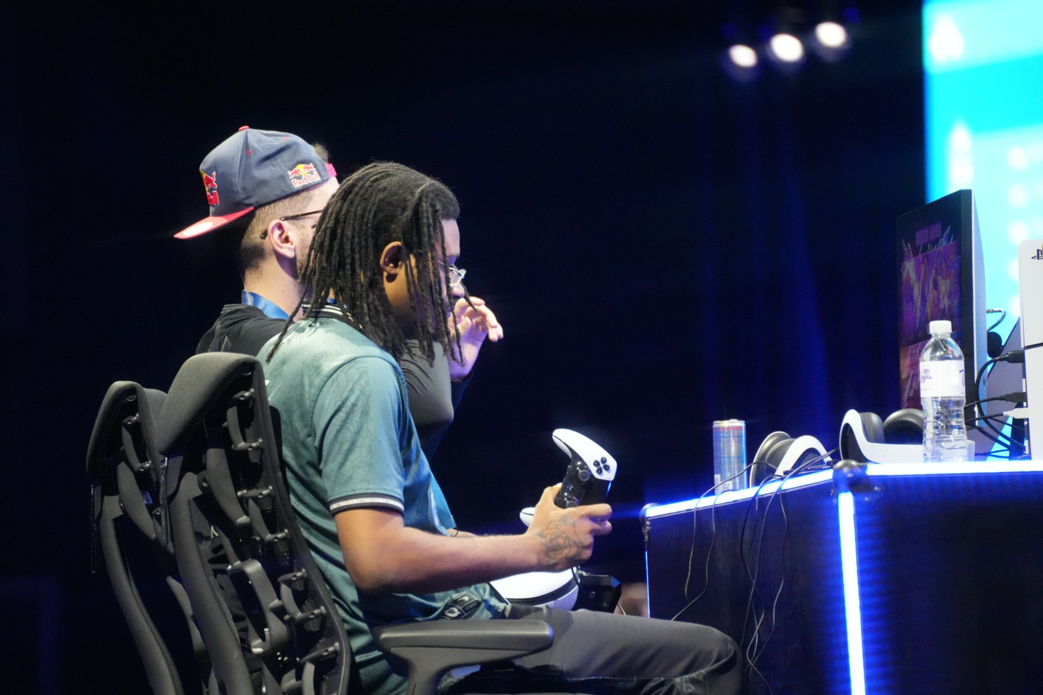 Two people sit at a table with computer monitors, one holding a game controller, in a dark room with bright lights.