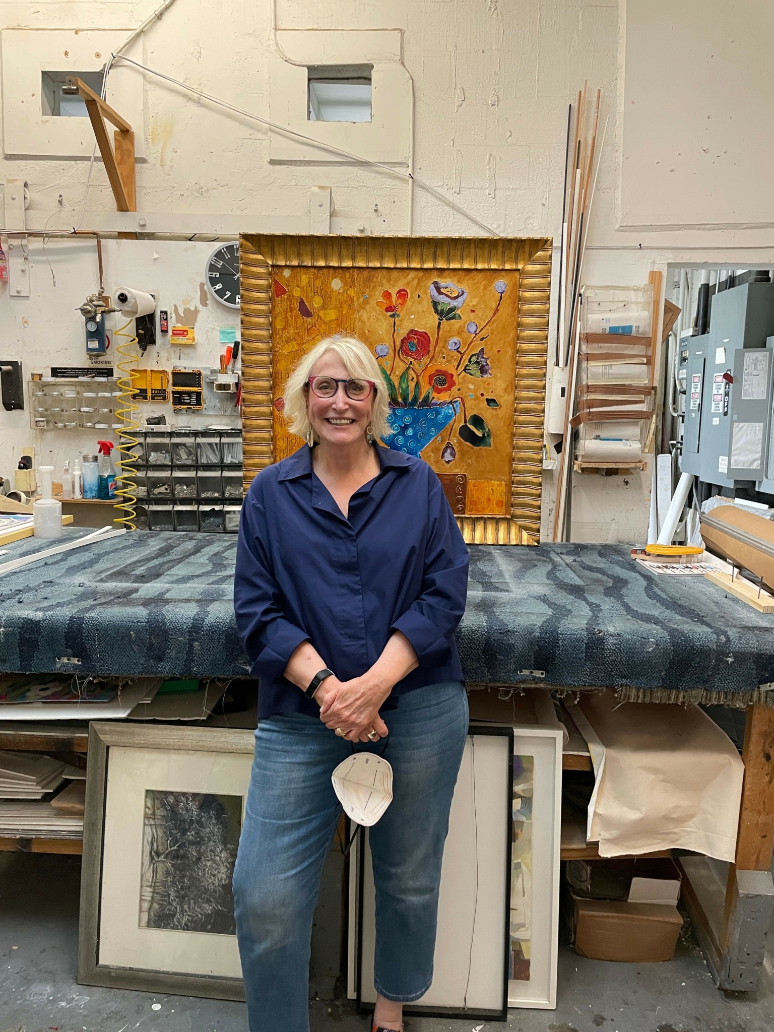 Woman with short blonde hair wearing a navy blue top and jeans, standing in a workshop or studio with tools and artwork.