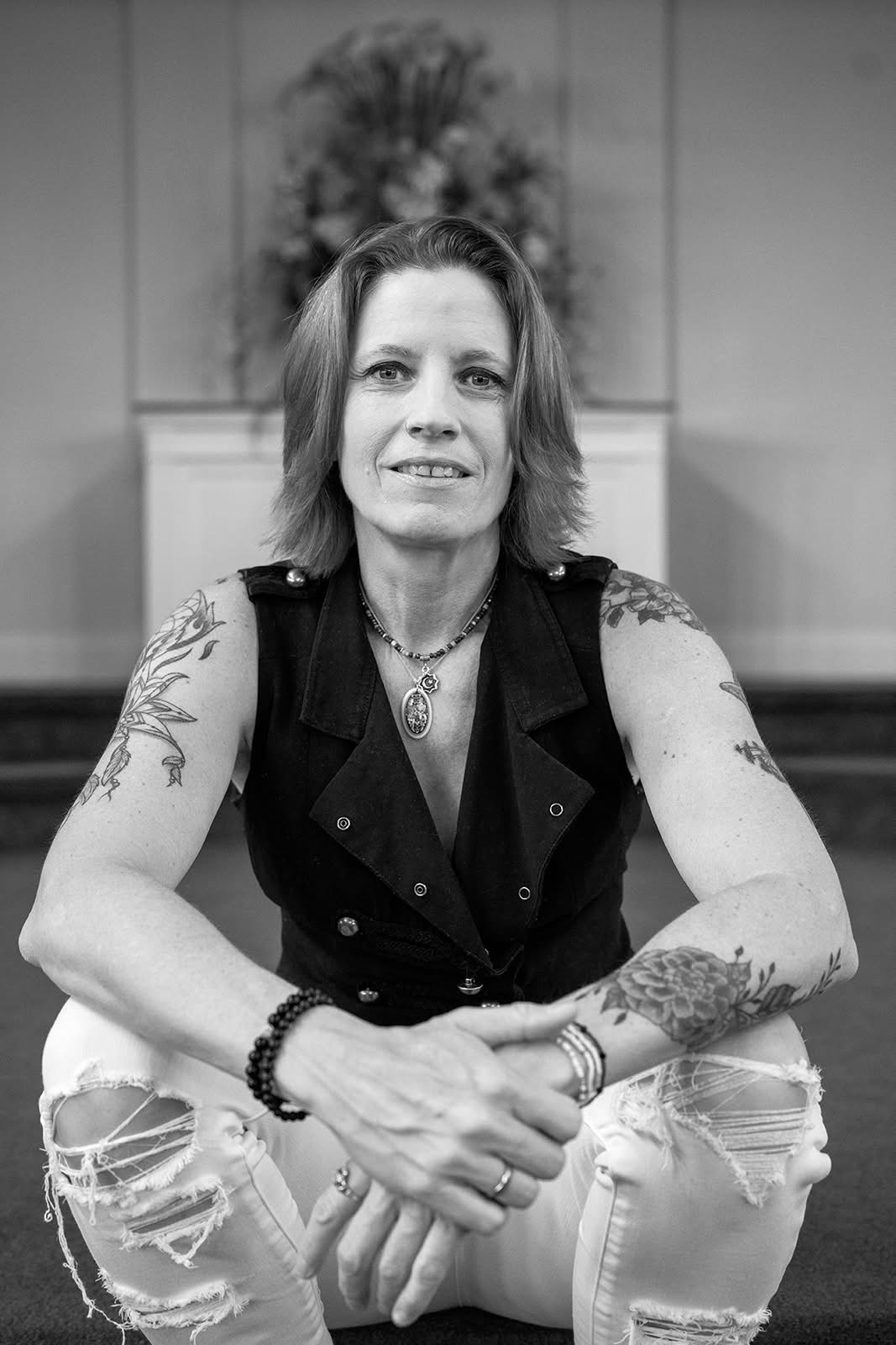 Woman with shoulder-length hair, tattoos on arms, wearing a sleeveless vest and ripped jeans, sitting with hands clasped.