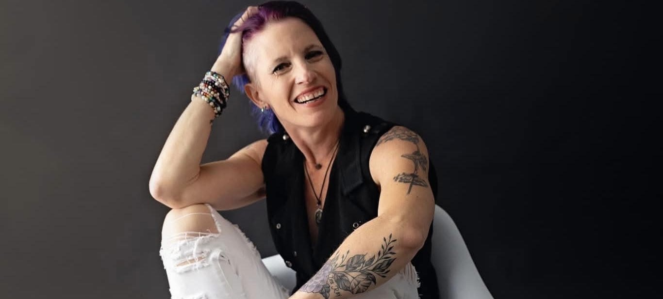 Woman with purple hair smiling, sitting with one arm resting on her head, tattoos visible on arms.