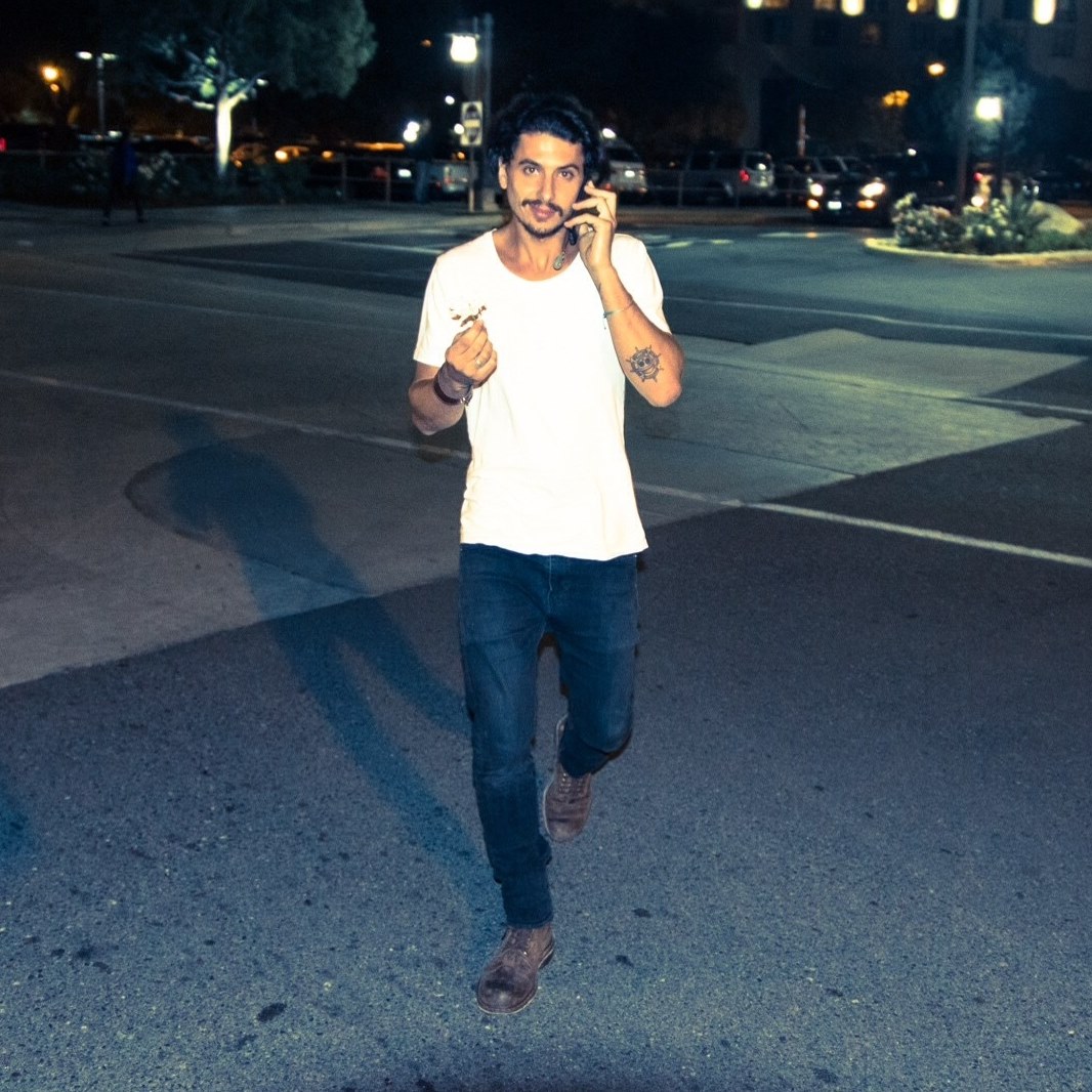 Young man walking on street at night, talking on phone, wearing white t-shirt and jeans.