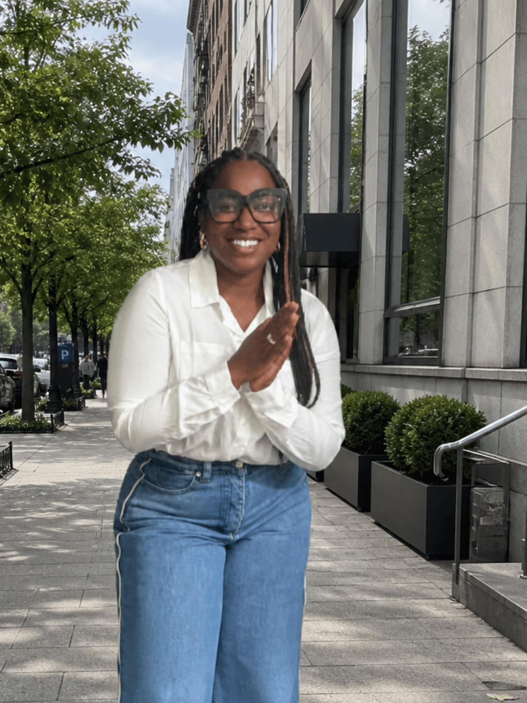 Woman with glasses smiling, standing outdoors on city sidewalk, trees and building in background.