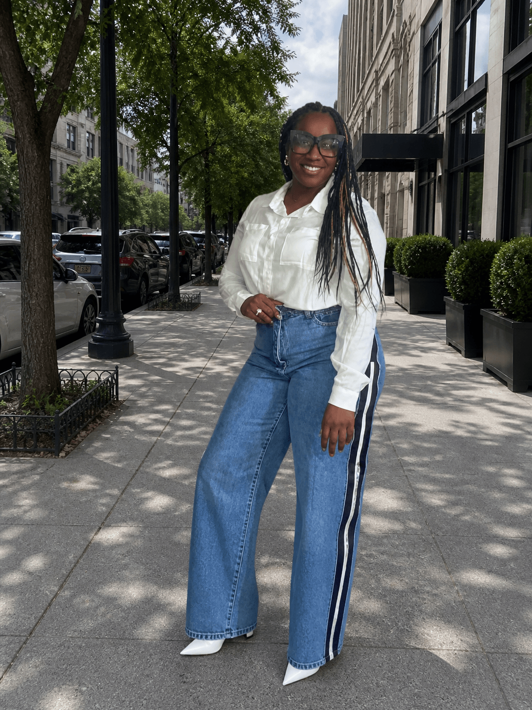 Woman with long braids and glasses standing on sidewalk in city street, smiling, wearing white blouse and wide-leg jeans.