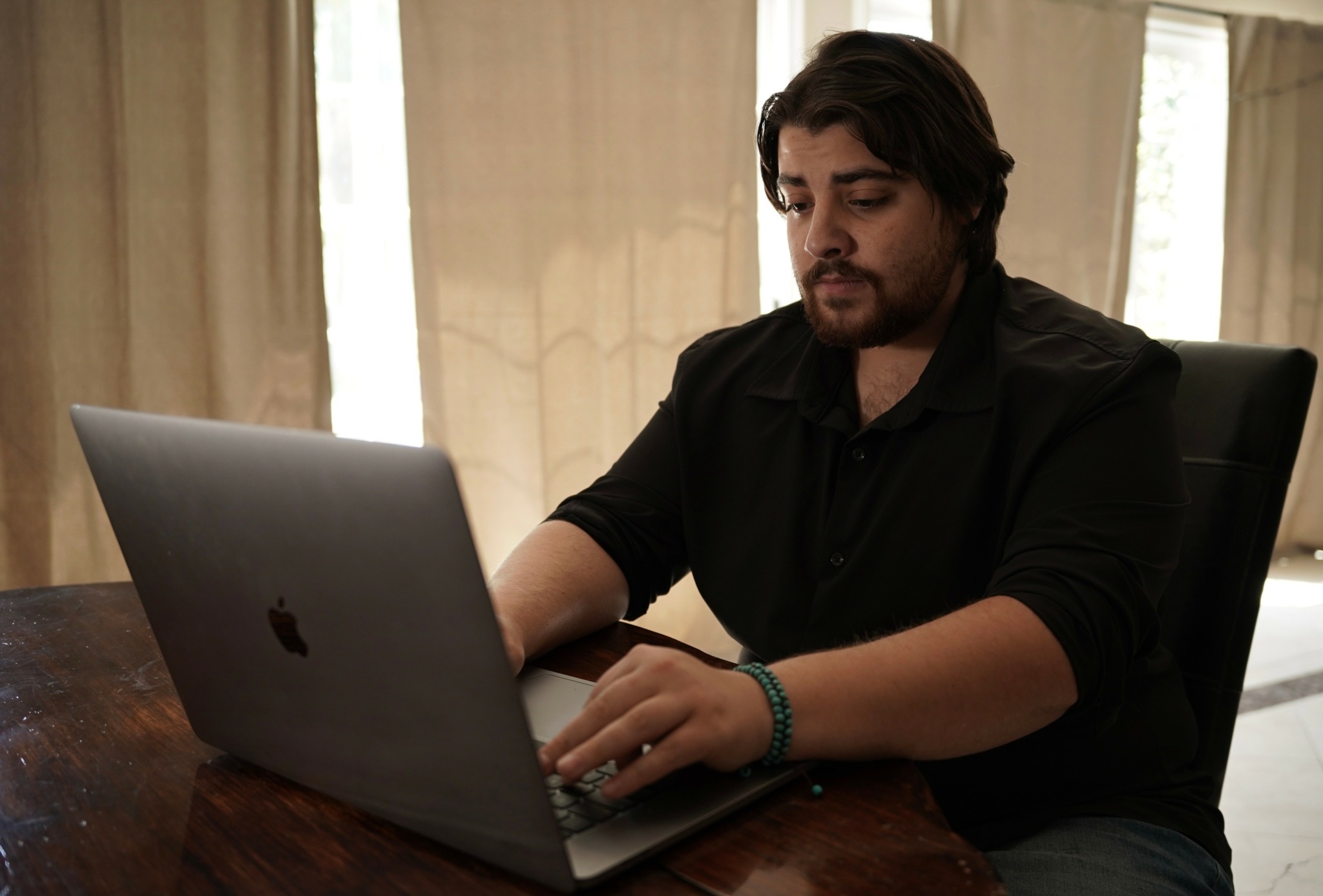 Man with dark hair and beard working on a laptop at a wooden table in a room with curtains.