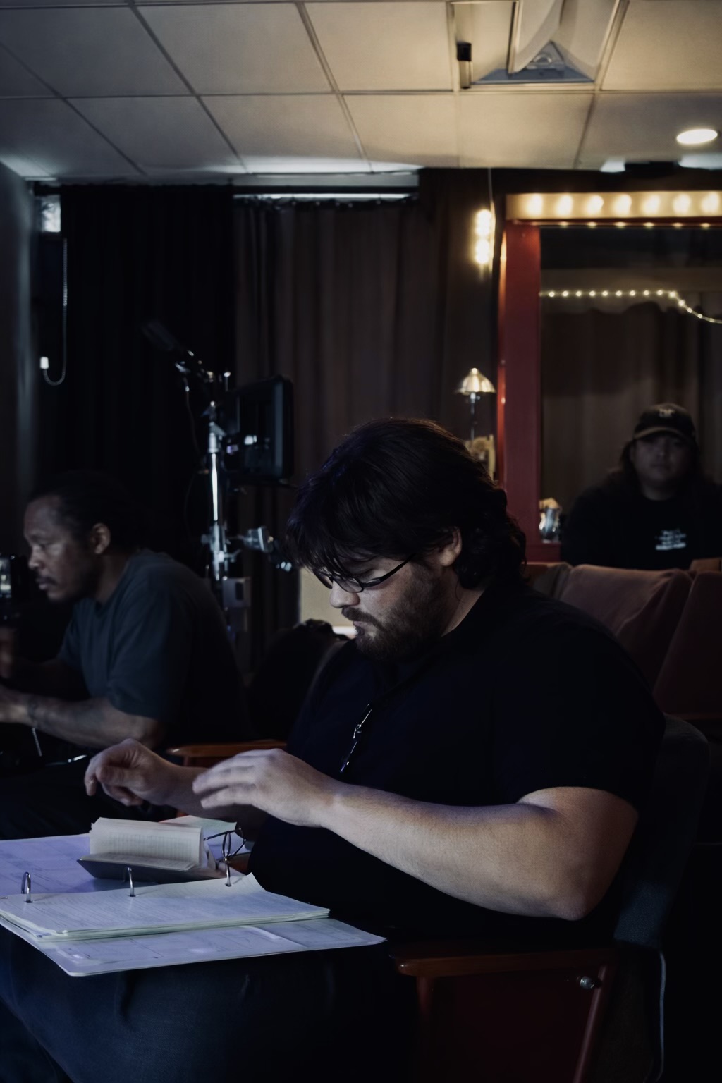 Person with glasses reading a book at a table in a dimly lit room with other people in the background.