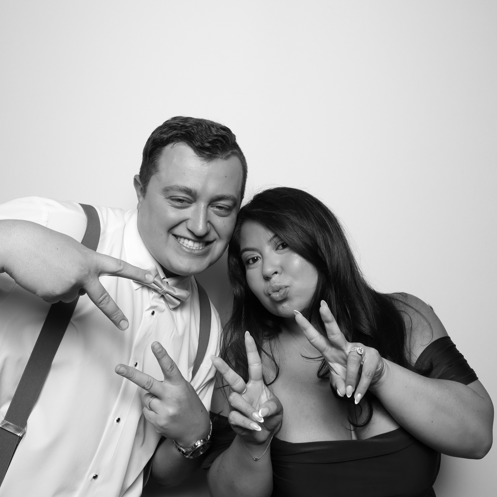 Two people smiling and making peace signs, posing closely together against a plain background.