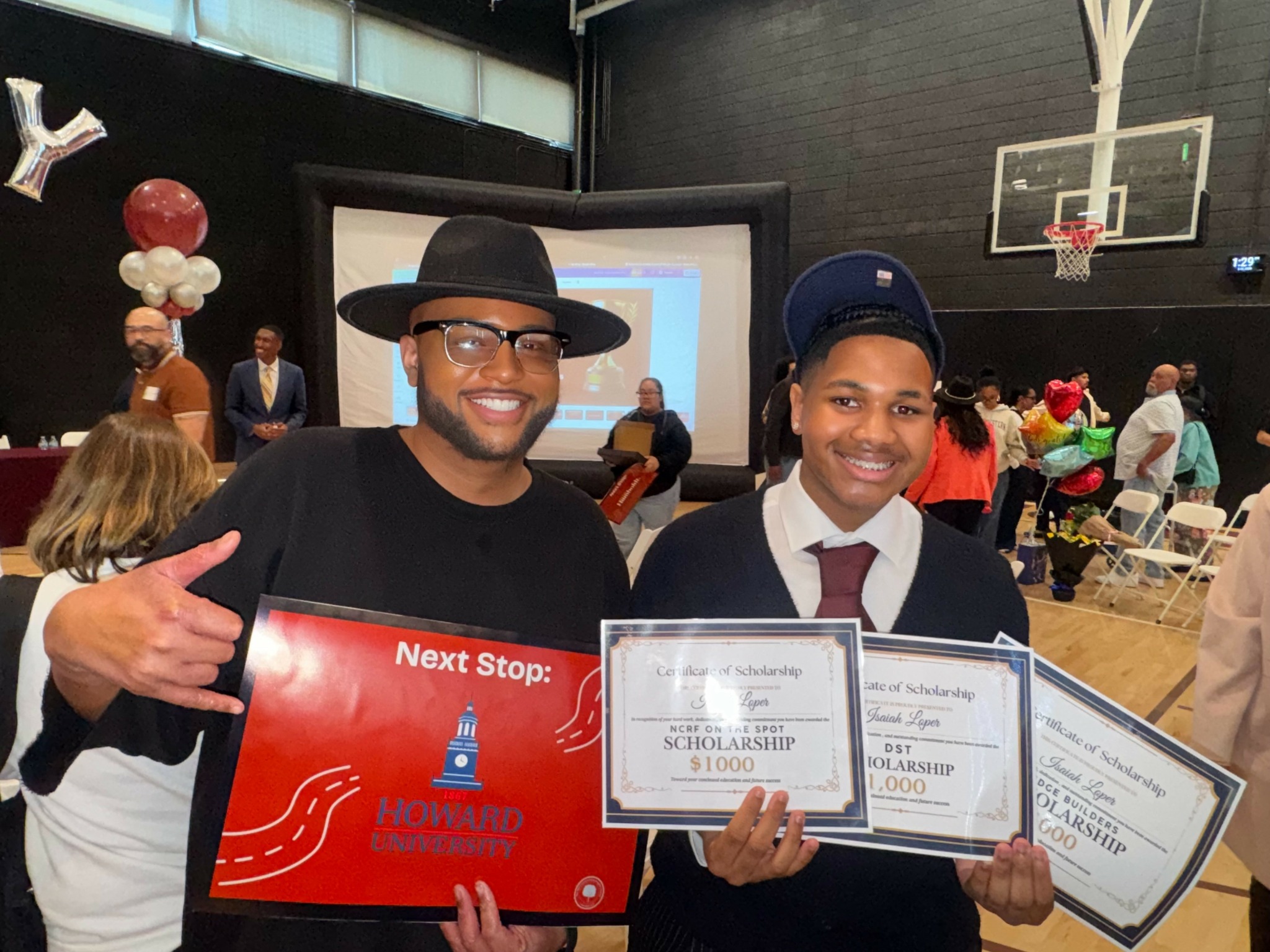 Two men smiling, holding a large check and certificate, in a gymnasium with people and a basketball hoop in background.