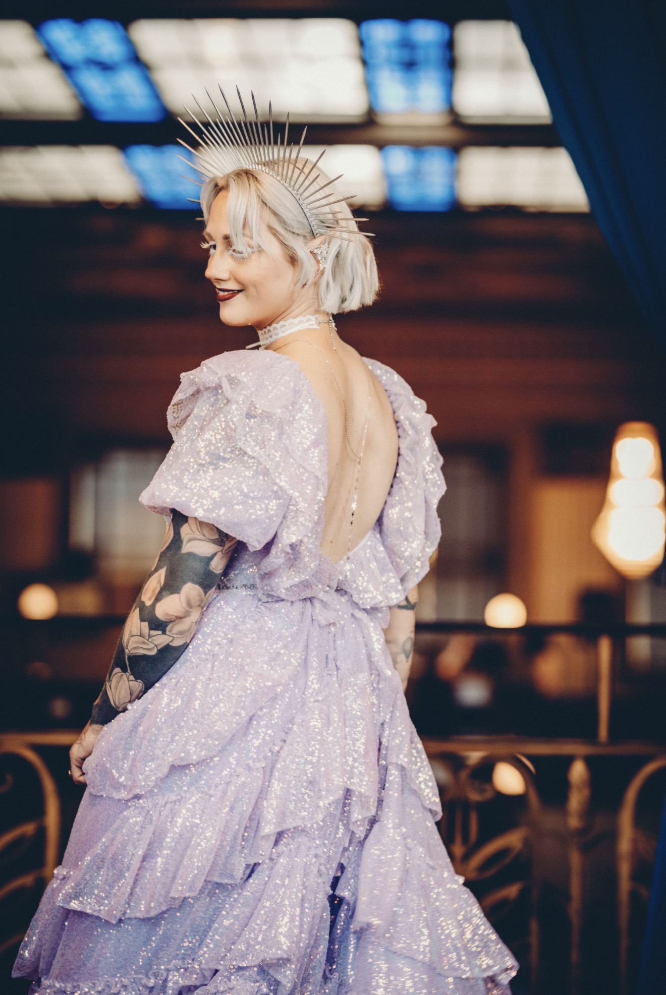 Woman in a lavender dress with ruffles, wearing a crown, smiling in an indoor setting with warm lighting.