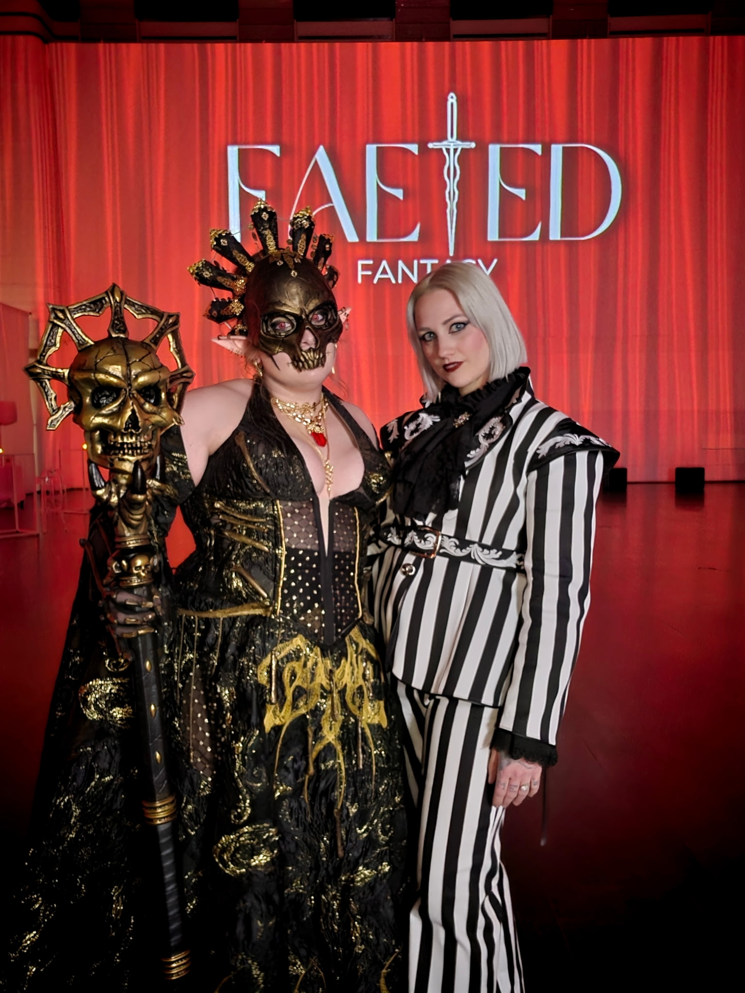 Two people in costumes pose in front of a red curtain with 'FAED' and 'FANT' text, one with skull makeup and black dress, the other with striped suit and white hair.