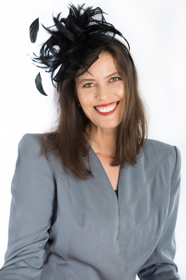 Woman with long brown hair wearing a gray blazer and a black feathered hat smiling.