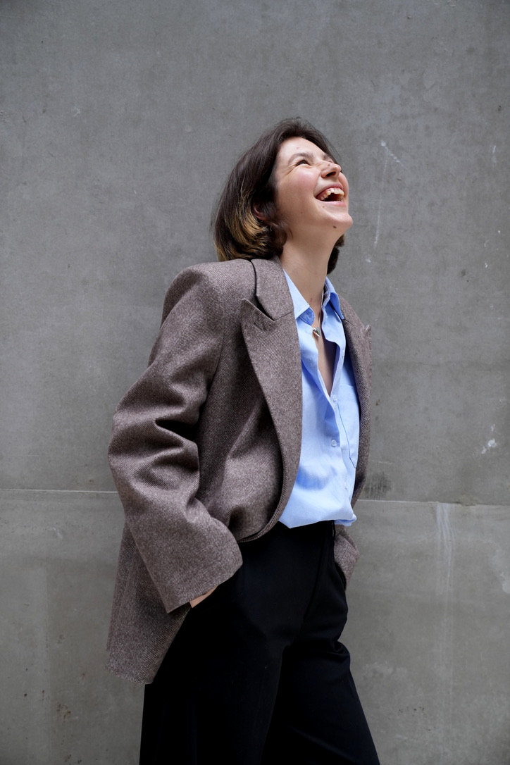 Woman laughing, wearing a blazer and shirt, standing against a plain wall.
