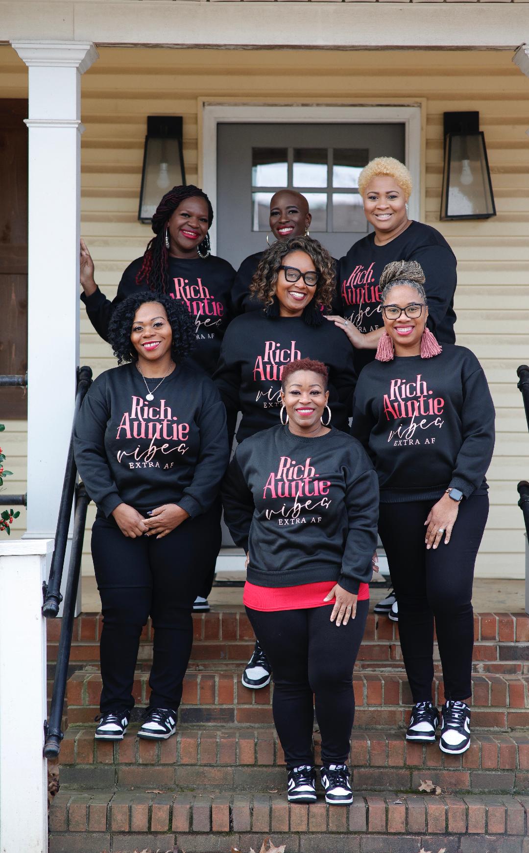 Seven women standing on front steps of a house, smiling, wearing matching black sweatshirts with pink and white text, some with glasses.